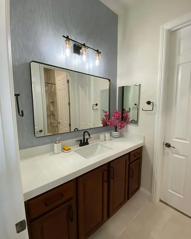 Bathroom vanity with a black-framed mirror, wall sconces and a textured blue-grey wallpaper